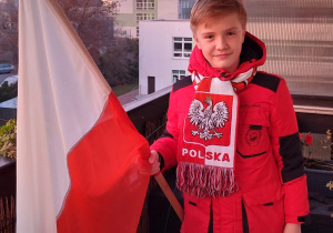 Chłopiec w czerwonej kurtce i szaliku z godłem Polski stoi na balkonie, a w ręku trzyma flagę biało-czerwoną..
